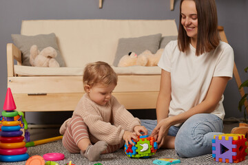 Photo of woman with dark hair sitting on floor, looking at her playing baby with smile, toddler girl studying new interesting toy, playing with colorful sorter.