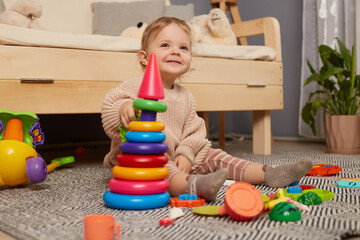 Indoor shot of cute smiling happy cheerful baby girl sitting on floor with her toys, playing with colorful pyramid, plays educational games alone at home.