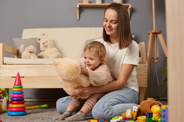 Indoor shot of dark haired woman wearing white T-shirt sitting on floor near sofa and holding her baby daughter, playing with soft toy teddy bear, expressing positiveness.