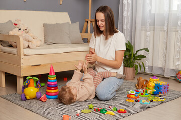 Indoor shot of young beautiful mother playing and having fun on the floor with her little daughter,...