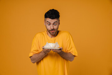 Excited hungry man holding cake and licking his lips isolated over yellow wall