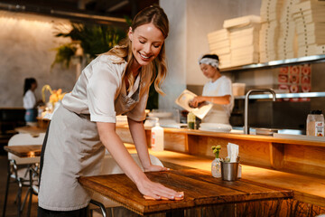 Young woman wearing apron cleaning table while working in restaurant - 561753076