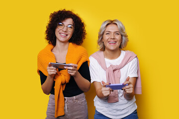 Two smiling young women gamer using smart mobile phone and playing games, isolated over yellow...