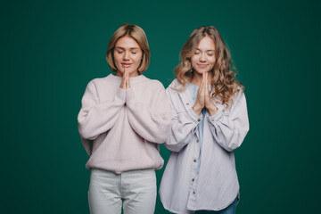 Two young women wearing casual clothes posing with palms together and eyes closed, ladies practicing yoga, standing against green wall. Happy lifestyle. People