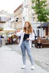 Obraz na płótnie Canvas Young redheaded beautiful woman walking through the old city and using her mobile phone for video blogging. Pretty smiling girl chatting online