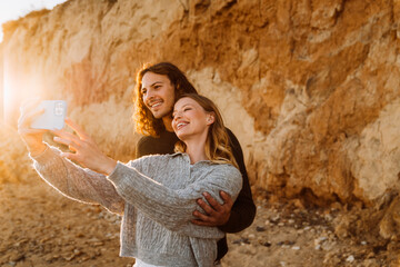 Beautiful young smiling couple wearing casual sweaters taking selfie while spending time at sunny beach