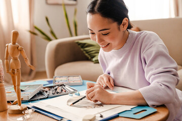 Smiling asian woman artist drawing with pencil while sitting at home
