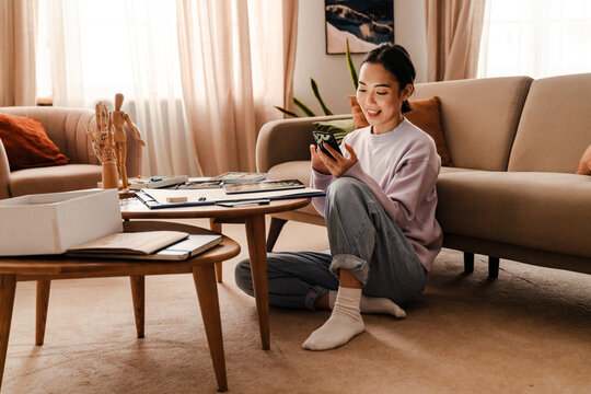 Smiling asian woman using mobile phone while sitting on floor at home