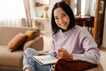 Fototapeta na wymiar Young smiling asian woman drawing on digital tablet with stylus while sitting in living room