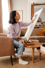 Young asian woman artist drawing on canvas while sitting on armchair at home