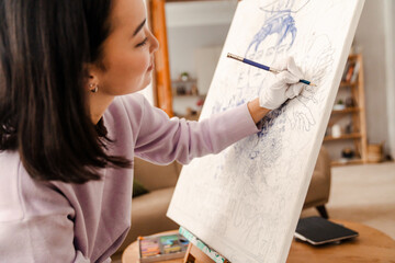 Young asian woman artist drawing on canvas while sitting on armchair at home