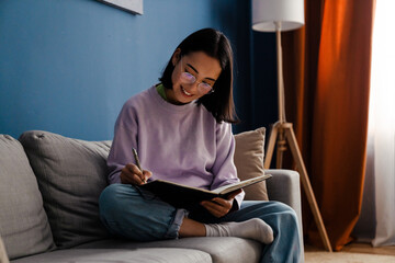 Beautiful asian woman smiling and writing down notes while sitting on sofa at home