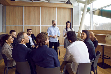 Business coach and psychologist talk to different people who are sitting in circle during group therapy session in office. Professional friendly male and female coaches train staff during teambuilding