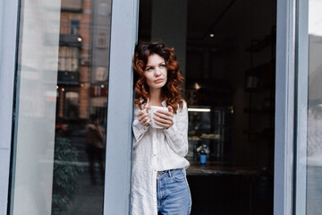 Stylish slim attractive woman with curly long hair wearing white shirt and jeans standing against modern cage with coffee and looking aside with happy smile . 