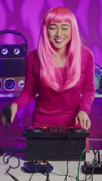 Vertical video: Musician with pink hair standing at dj table dancing and having fun while mixing stereo sounds with electronics. Smiling artist enjoying to perform music using professional mixer