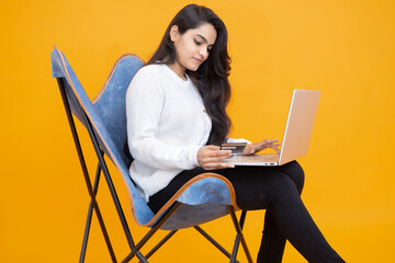 Young indian girl using credit card for online shopping on her laptop while sitting on chair isolated over orange yellow background. Studio Shot, Copy space, Asian woman using computer.