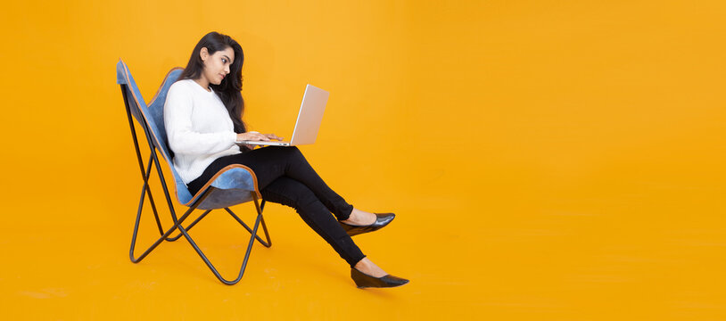 Young indian girl wearing white T-shirt using laptop while sitting on chair isolated over orange yellow background. Studio Shot, Copy space, Asian woman using computer.Technology concept.