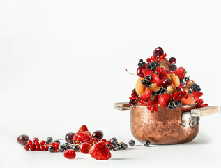 Copper cooking pot full with various summer berries and fruits on white background, front view
