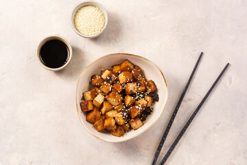 Sticky tofu - vegan soybean protein source in soy sauce and sesame oil marinade roasted on a pan sprinkled with sesame seeds, in a beige colored bowl on a concrete background, top view