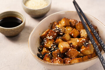 Sticky tofu - vegan soybean protein source in soy sauce and sesame oil marinade roasted on a pan sprinkled with sesame seeds, in a beige colored bowl on a concrete background