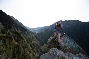 a beautiful girl in a dress sits on a large stone in the mountains. hipster in black shoes on the background of mountains. a woman in a dress. general plan