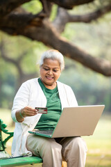 Indian senior woman using laptop and bank card at park.