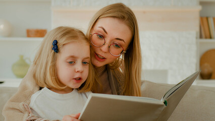Fototapeta na wymiar Caucasian mother in eyeglasses with little small cute daughter preschool child girl baby reading book in living room at home mom with kid read literature at couch sofa elementary learning education