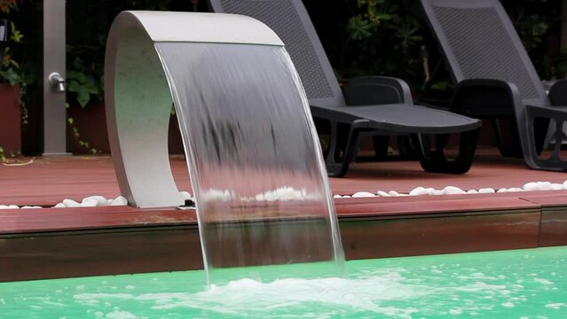 Stainless steel swimming pool wall waterfall jet on luxury resort. Water feature fountain with clean green water falling down under high pressure. Lounge area with sun loungers by a pool in summer.
