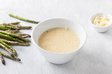 White ceramic bowl of beaten eggs with milk, cheese and fresh asparagus shoots on a light gray background. Cooking a delicious homemade omelet