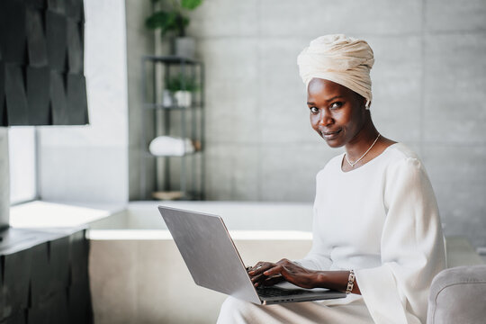 Purposeful African woman in white turban works at home using laptop looks at camera. Confident Brazilian businesswoman remote managing company. American Female Writer typing text on computer at hotel.