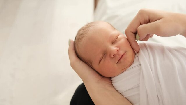 Mother hands strocking head and cheak of newborn baby swaddled in white fabric. Girl calming sleeping on her legs infant. Little child napping and his mom caring about kid