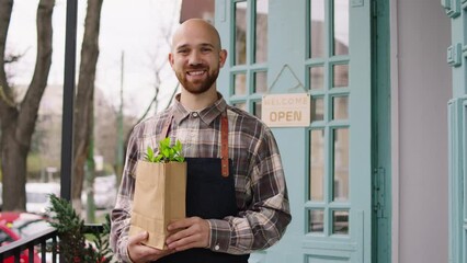 Cute floral shop owner and florist man smiling large in front of the camera while holding a eco bag with some plants inside beside of the shop