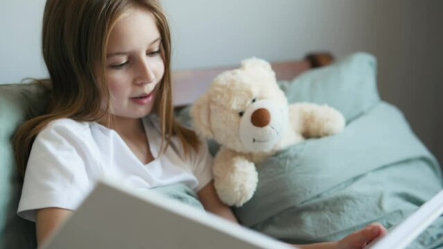 Cute girl reading book fairy tale to teddy bear in bed. Preteen female kid with toy and album at home