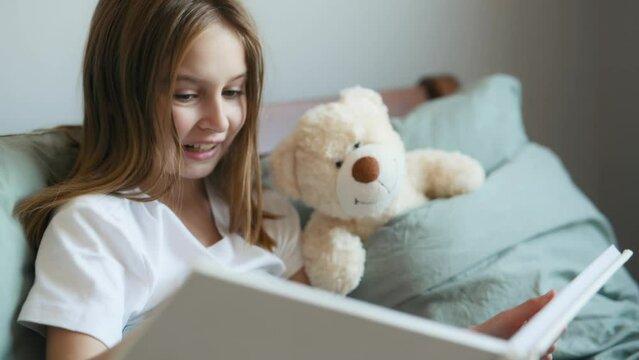 Pretty little girl reading book to teddy bear in bed. Preteen female kid with toy and album at home