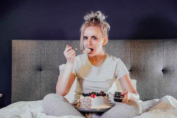 Emotionally stressed, shocked woman eating cake and holding a remote control while watching TV in...