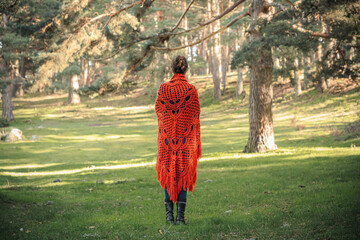 A woman in a red knitted shawl stands in a forest clearing and looks ahead.