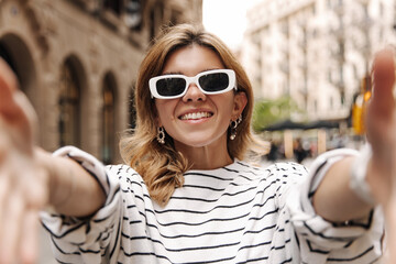 Cheerful european stylish woman reach hands to camera on the blur background. Fair-skinned blonde in sunglasses smiling. Emotional concept