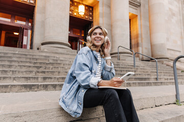 Fototapeta na wymiar View of attractive smiling woman looking away keep hands on headphones. European young student in hands phone, sitting at the street. Concept of leisure life