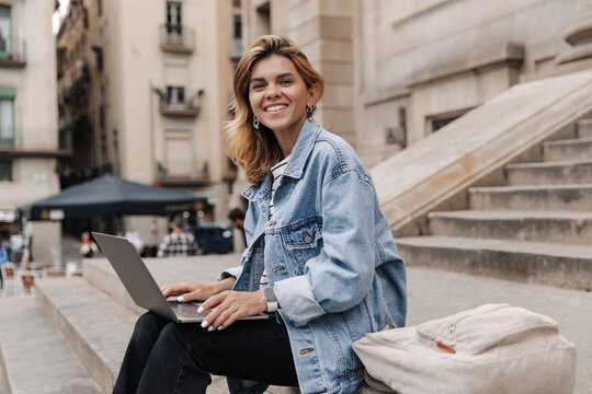 Caucasian charming smiling woman working outdoors on laptop. Wearing blue jacket, black pants. lying near backpack sitting on the stairs. Use technology concept