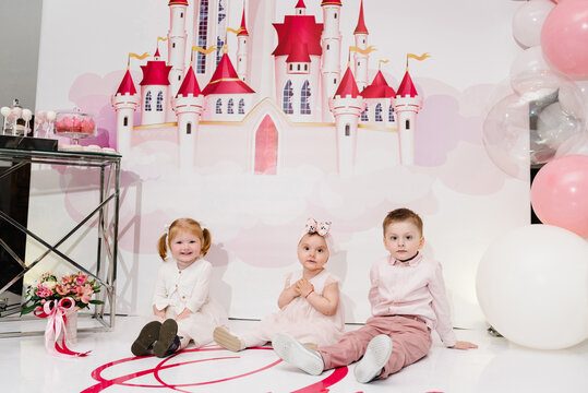 Portrait of a boy and girls little luxury princess in dress on background pink balloons and castle. Concept of birthday celebration. A childs, kids sitting on the floor and playing.