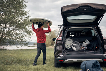 Windsurfer and camper unpacking equipment from a car in nature near the lake shore.
