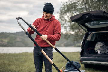 Windsurfer and camper unpacking equipment from a car in nature near the lake shore.