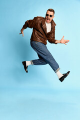 Fototapeta premium Portrait of brutal, handsome man in jeans, leather jacket and sunglasses posing, jumping over blue studio background. Concept of emotions, lifestyle