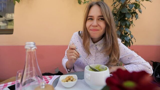beautiful blonde girl eats green soup with smiles on the summer terrace in the restaurant. a woman is happily having lunch at a table with a white plate. middle plan