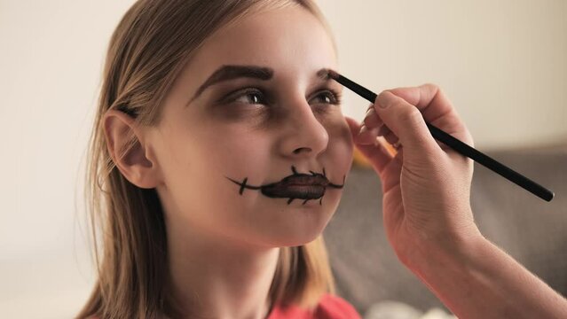 Mother making Halloween makeup to preteen girl daughter and painting spooky black lines on face. Pretty child with creepy cosmetics at October holiday death day celebration