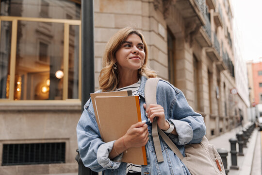 Picture of pretty fair-skinned woman walking with notebook on the street. Caucasian student looking away holding backpack. Cite style concept