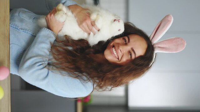 Vertical video. Portrait of the smilling woman holding the fluffy easter rabbit, wearing bunny ears. Smiling lady is getting ready for the spring holiday. Preparation for easter