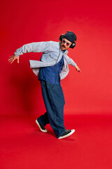 Fototapeta na wymiar Hip hop. Portrait of man in stylish clothes, oversized jeans, shirt and hat posing, dancing over red background. Concept of modern fashion, lifestyle, music culture