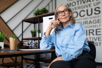 a mature businesswoman with a blue strict shirt sits against the backdrop of an office in glasses...