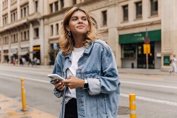 View of attractive caucasian woman loking away and smiling in the city. Stylish lady holding phone in hands staying on street. Lifestyle, street style concept